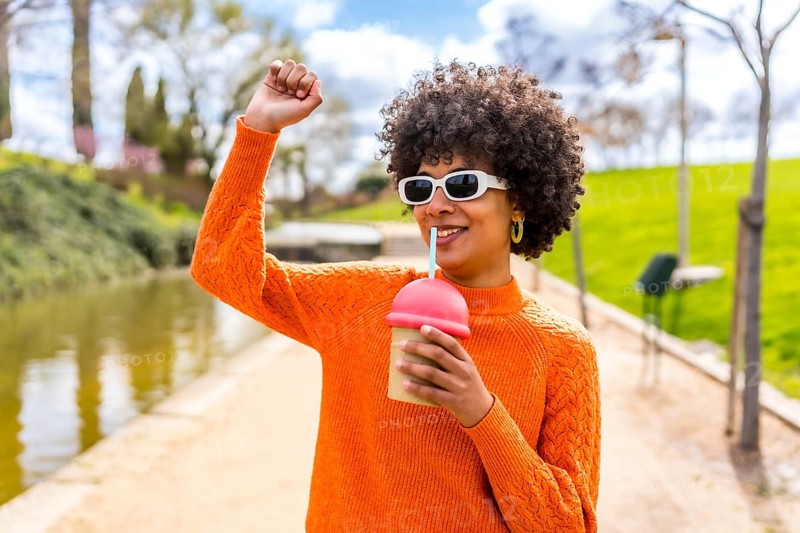 Happy young woman with curly hair and orange sweater, enjoying a smoothie in a park by the river, raising her arm in a carefree gesture
