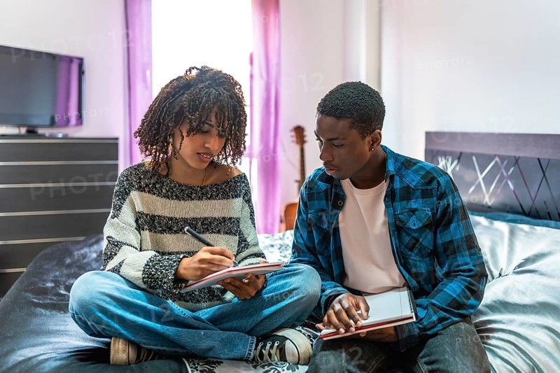 Two focused college students are sitting on a bed, sharing notes and preparing for exams in their dorm room