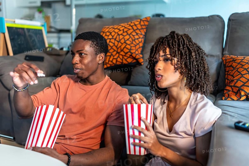 Relaxed couple enjoying a movie night at home, sitting on the floor in front of their sofa and eating popcorn from striped buckets