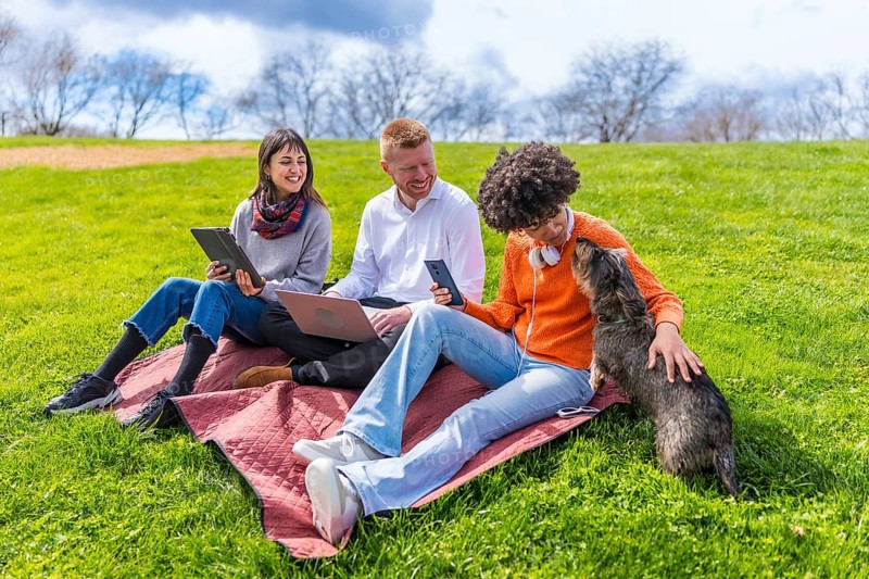 Three young coworkers are taking a break from work outdoors, using laptop, tablet and smartphone, while petting a dog in a park