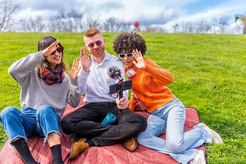 Three content creators waving cheerfully at the camera while filming a video blog outdoors in a sunny park, enjoying their time together