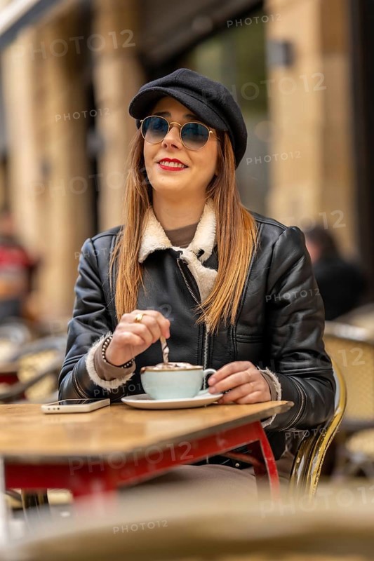 Young woman wearing sunglasses and hat stirring cappuccino at a parisian outdoor cafe, enjoying a relaxing moment