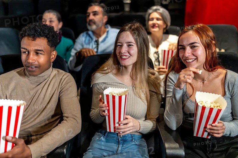 Happy multi ethnic friends watching film together and eating popcorn, having fun at the movie theatre