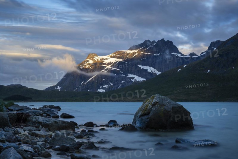 Landscape with sea and mountains on the Lofoten Islands, view over the fjord Flakstadpollen to mountains. Rocks in the foreground. At night at the time of the midnight sun, some clouds in the sky, the sun shining on the mountain. Long exposure. Early summer. Flakstadoya, Lofoten, Norway, Europe