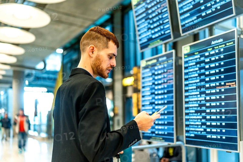 Businessman checking flight information on an airport display while using a smartphone, staying connected during travel and planning his journey