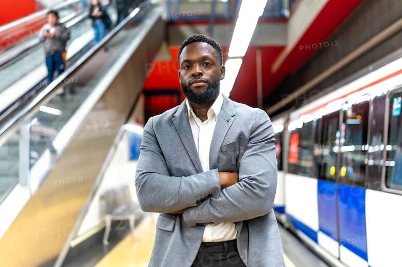 Young black businessman standing with crossed arms in subway station, waiting for transportation, commuting to work