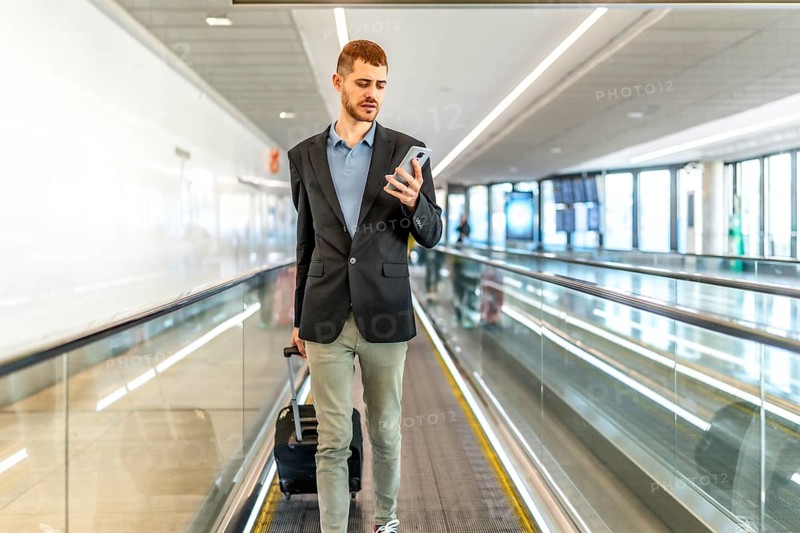 Businessman pulling suitcase and using smartphone while walking on moving walkway at airport