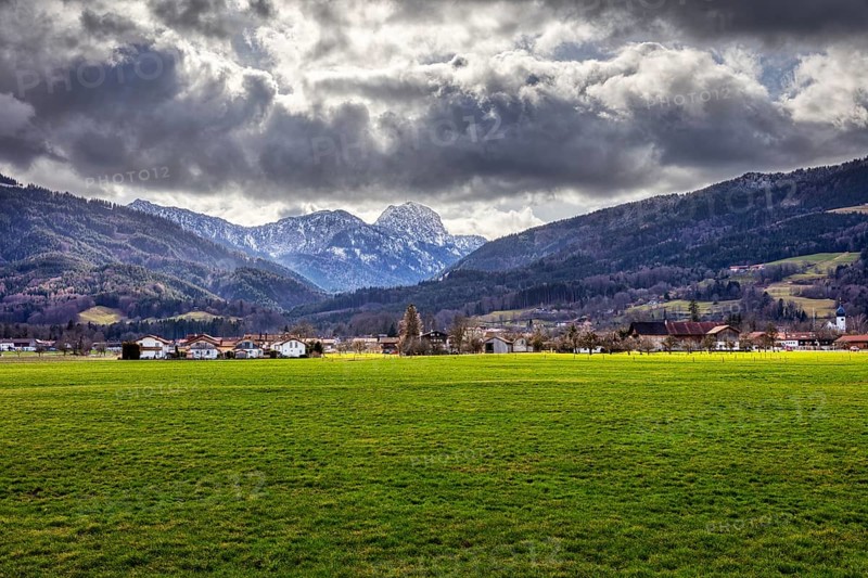 Dramatic Alpine landscape with green meadow in the foreground and cloudy sky over a village, Bad Feilnbach