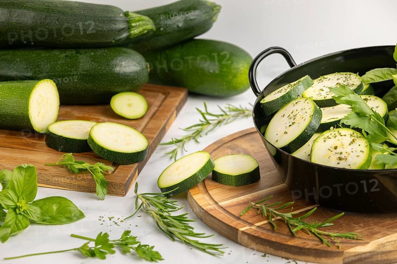 Sliced and seasoned courgettes with fresh herbs in a black pot, next to stacked courgettes on a wooden board