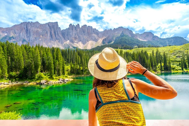 Woman wearing straw hat admiring lake carezza with latemar mountain range in background, dolomites, italy