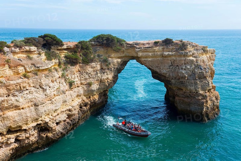 A boat with tourists sails through a natural rock arch over the clear blue sea, Arco de Albandeira, Lagoa, Algarve, Portugal, Europe