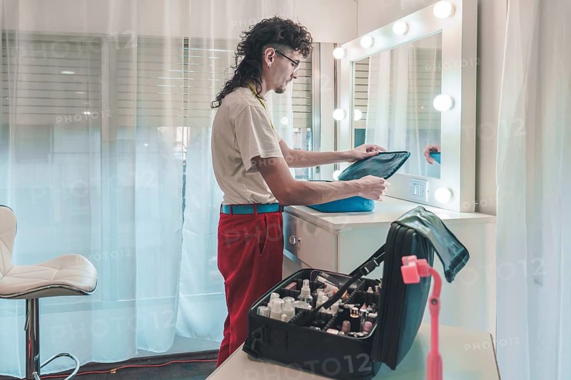 Professional makeup artist unpacking cosmetics bag in brightly lit dressing room, preparing for work with various beauty products and tools on the table