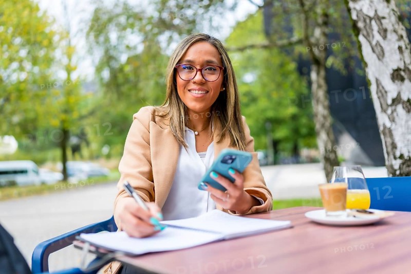 Latin woman using phone while writing notes drinking coffee sitting on a tranquil terrace of a cafeteria in the city