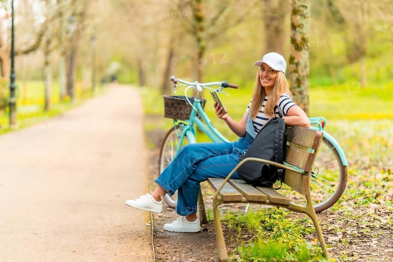 Happy tourist is using her smartphone while resting on a bench in a park with her bicycle next to her