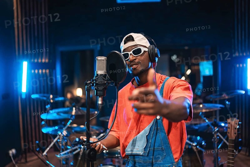 Talented musician recording vocals, pointing at you, wearing trendy sunglasses and cap, creating new music in a modern studio