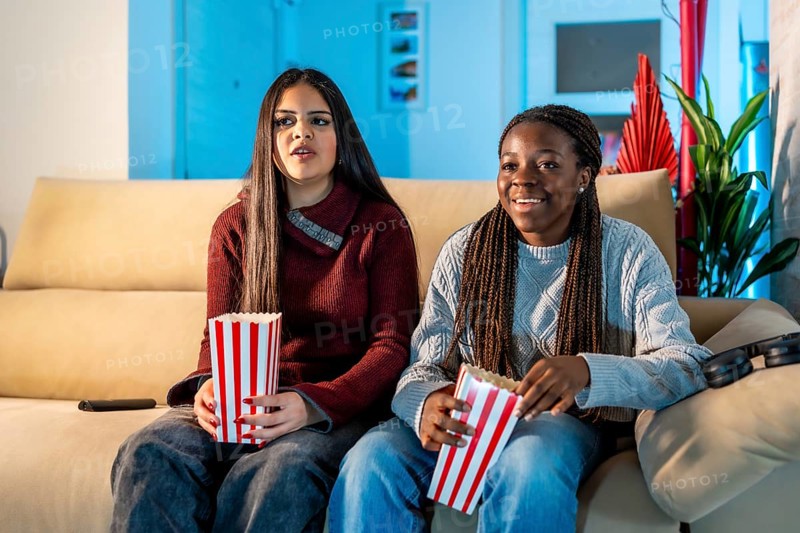 Two teenage girls enjoying a movie night at home, sitting on a comfortable sofa, eating popcorn from striped boxes, and having a great time together