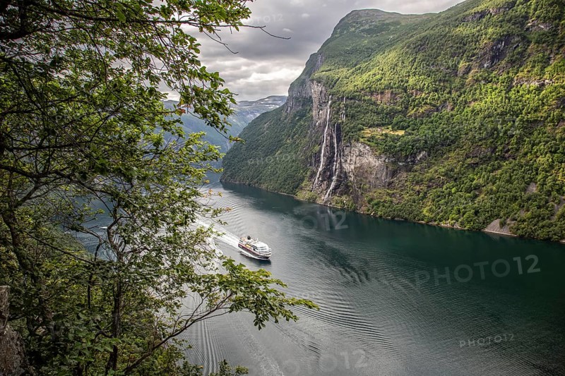 A cruise ship sails through the Gairanger Fjord surrounded by steep mountains and the Seven Sisters waterfall in Norway Europe