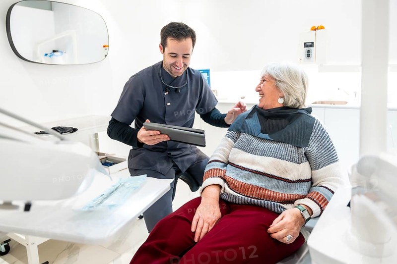 Dentist showing something on tablet to smiling senior woman sitting in dentist chair, during consultation in dental clinic