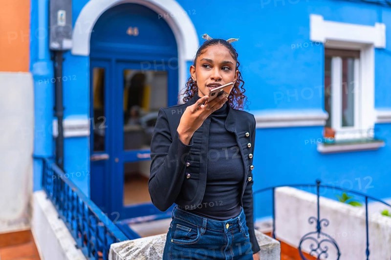 African american woman sending a voice message with smartphone next to colorful blue house