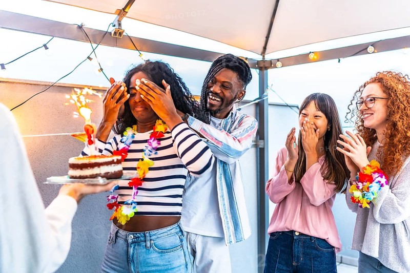 African young woman with eyes covered receiving a birthday cake from multi-ethnic friends at terrace