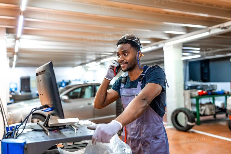 Young mechanic wearing gray uniform and protective glasses using computer and talking on phone while working in car service