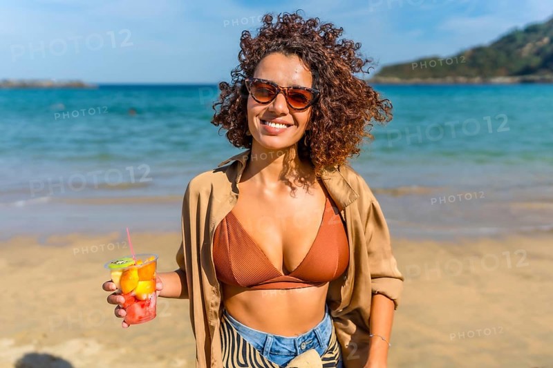 Confident arab woman holding a glass of tropical fruit at beach under the sun in a windy day