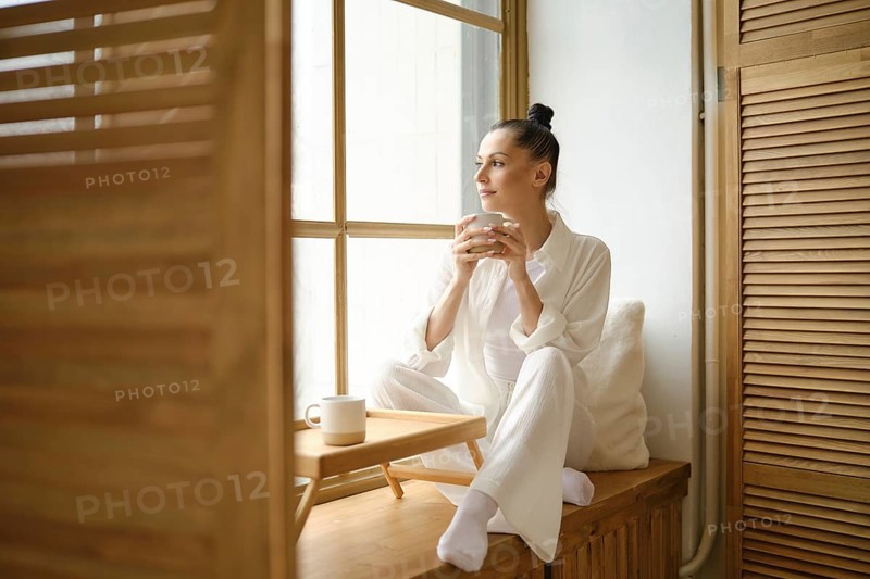 Dreamy woman in white clothes sitting on windowsill and holding cup of coffee or tea with both hands