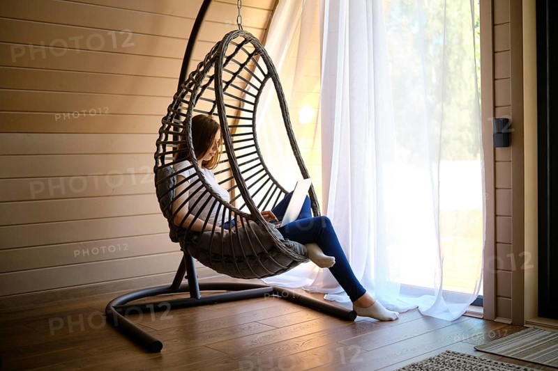 On a hot sunny day, a young woman sits in a hanging swing chair with a laptop in an air-conditioned room of log cabin