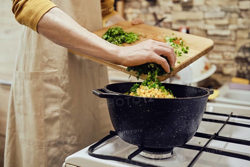 Unrecognizable man pours chopped green onions into a saucepan on a gas stove