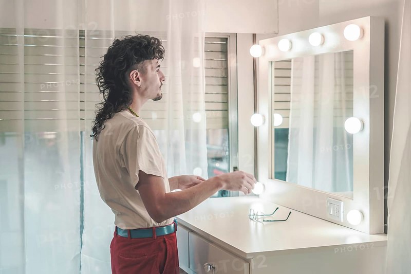 Young man with a mullet hairstyle standing in front of a lit mirror, holding items, dressed in a white t-shirt and red pants, preparing for the day in a cozy bedroom