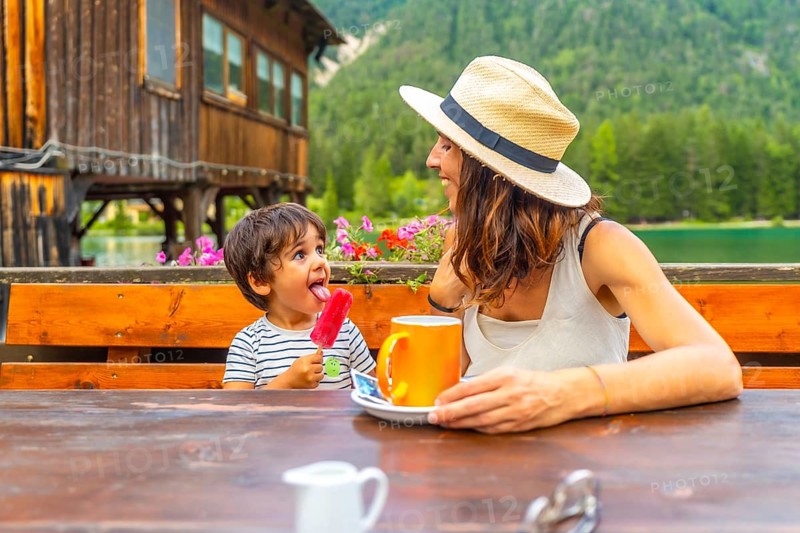 Mother and son enjoying a lakeside snack with a colorful popsicle and warm beverage against a stunning mountain backdrop