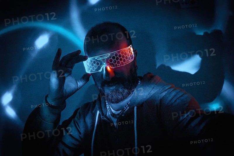 Bearded man adjusting futuristic glowing smart glasses in dark cyberpunk setting with blue and turquoise neon lights