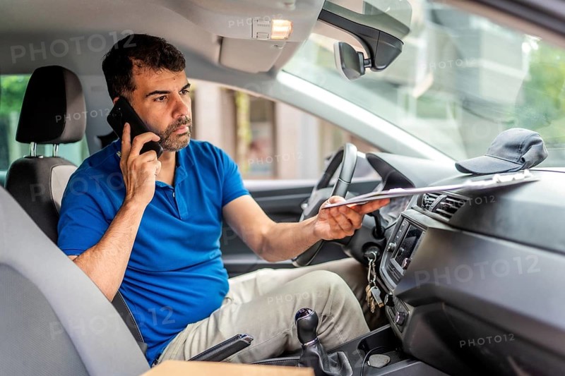 Mature caucasian male delivery person talking to the mobile phone sitting inside a van