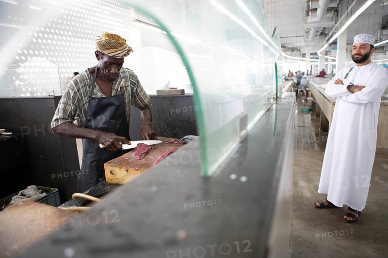 Omani man filleting a fish in Muttrah fish market, an Omani man in dishdasha watches, Muscat, Oman, Asia