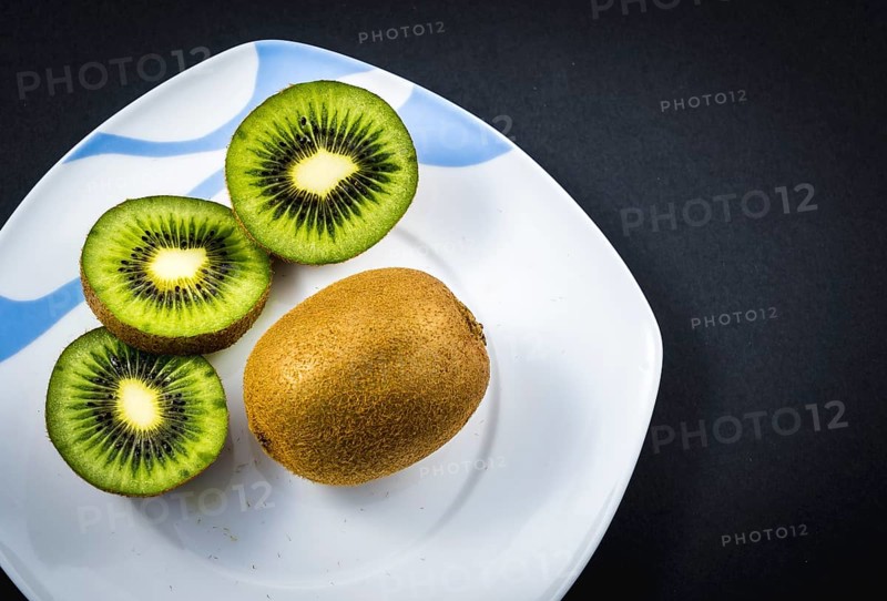One whole kiwi and two cut kiwis on a white plate with a black background