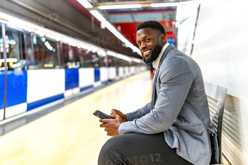 Young black businessman sitting on bench in subway station using smartphone while waiting for train
