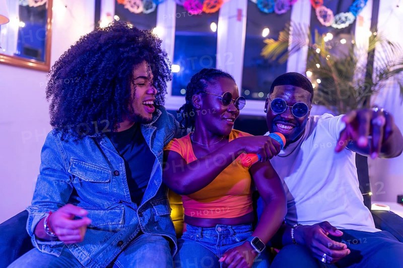 Happy people singing karaoke during night party sitting on a sofa and laughing