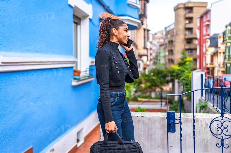 Side view photo of an african american casual businesswoman walking talking to the mobile among colorful street carrying a laptop bag