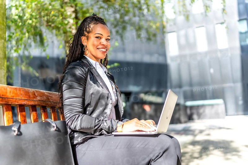 Side view portrait of a successful latin businesswoman working with laptop sitting on city bench outdoors in a sunny day