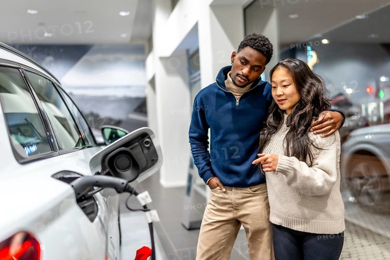 Multiethnic couple examining charging port of electric vehicle at car dealership, embracing sustainable transportation
