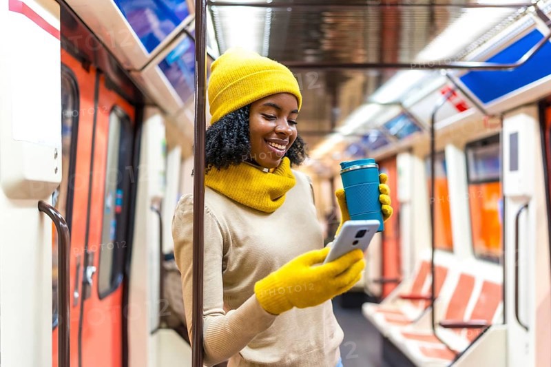 Smiling woman wearing yellow winter clothes using phone and holding reusable coffee cup while commuting on public transport