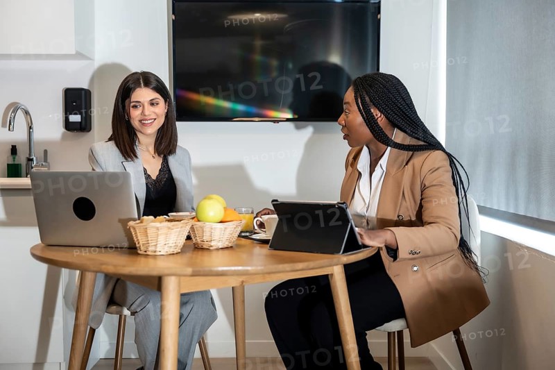 Two businesswomen are enjoying breakfast and working with laptop and tablet in their hotel room during a business trip
