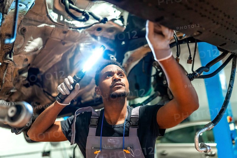Young mechanic using flashlight, inspecting underside of vehicle raised on lift in professional garage