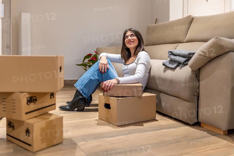 Young woman taking a break from unpacking, sitting on the floor of her new apartment and looking up dreamily