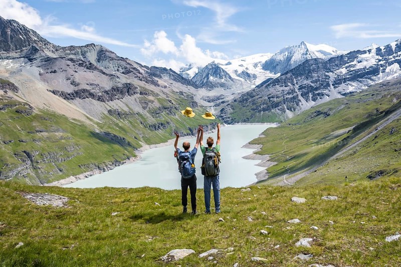 Two friends throwing yellow hats in the air, mountaineers in front of a blue mountain lake, Lac des Dix reservoir, behind mountain peak Mont Blanc de Cheilon with glacier, Hérménence, Valais Alps, Valais, Switzerland, Europe