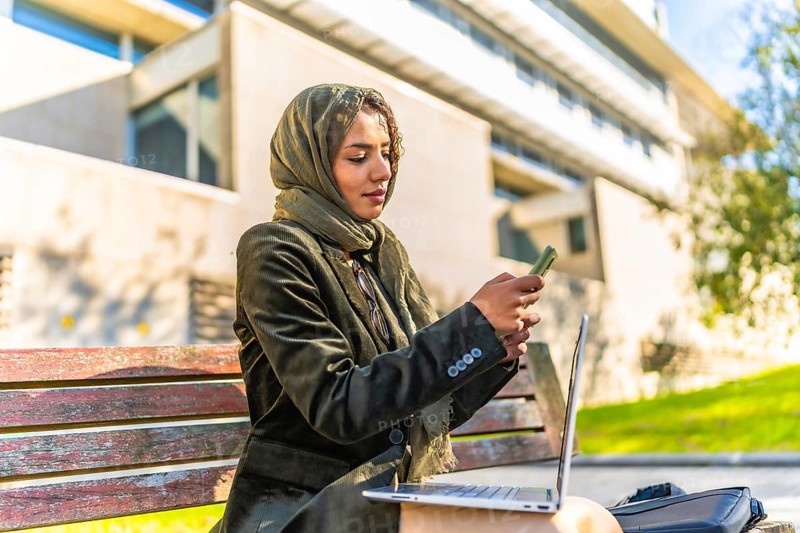 Muslim businesswoman using mobile working on remote with a laptop sitting on a bench in a park