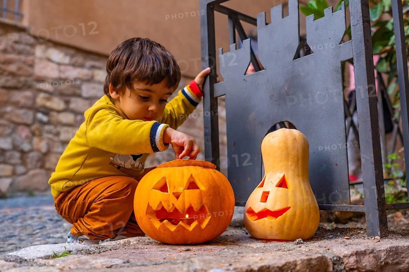 A young boy is playing with two pumpkins, one of which has a scary face. The scene is set in a courtyard, with a stone wall in the background. The boy is wearing a yellow shirt and brown pants