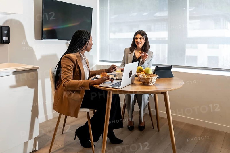 Two businesswomen are enjoying breakfast and working on a laptop during their business trip, showcasing the blend of work and leisure in a hotel environment
