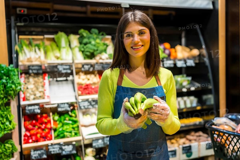 Young female greengrocer wearing gloves holding a bunch of organic bananas, smiling in a supermarket with fresh produce