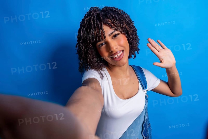 Smiling young woman with curly hair, taking a selfie and waving her hand against a vibrant blue background, radiating happiness and friendliness